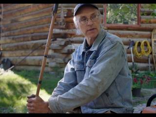 muž sedí před srubem s tyčí v ruce a nářadím vedle sebe